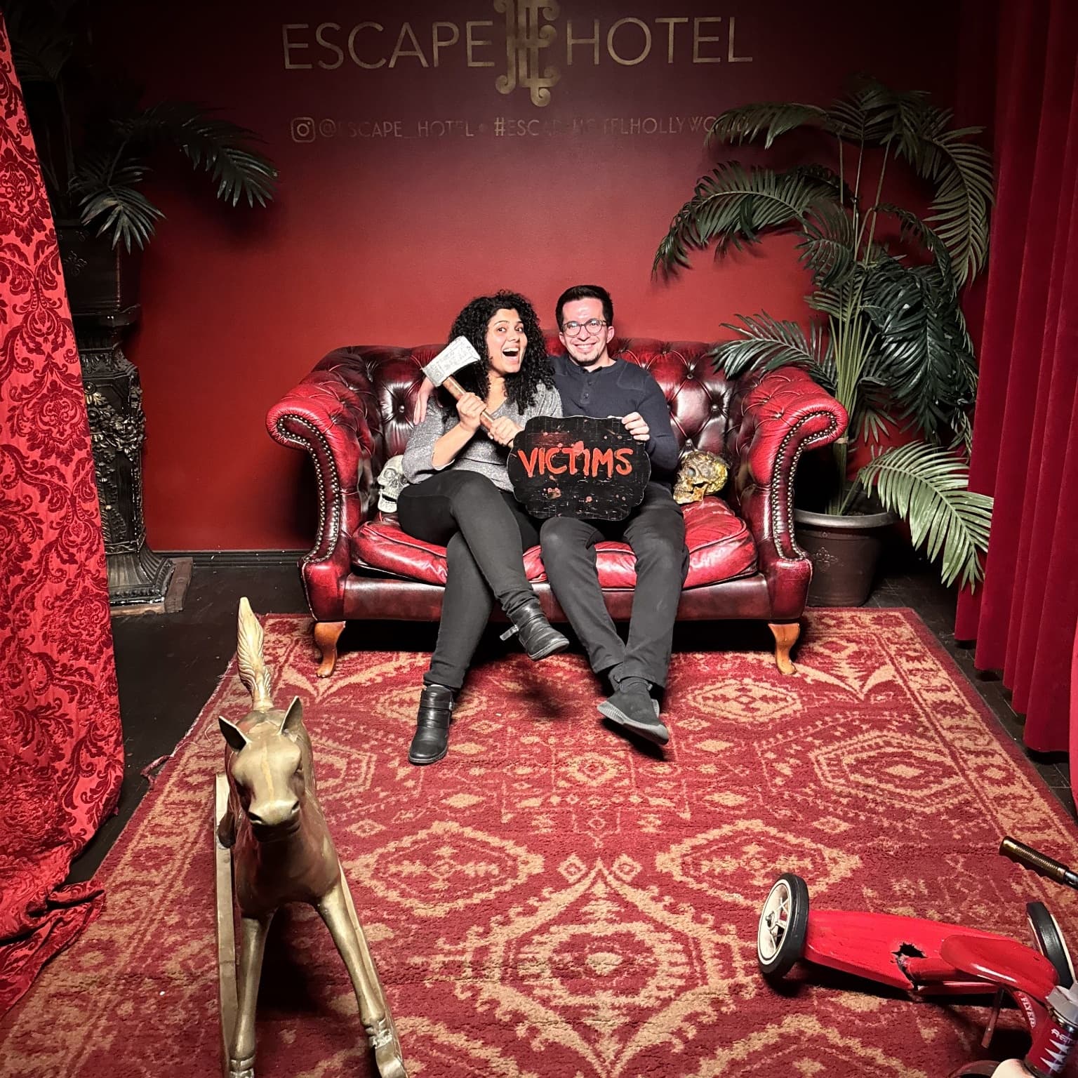 Gayatri holding a prop axe and Oleg holding a VICTIMS sign on a red leather couch at the Escape Hotel